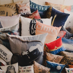 May include: A collection of decorative throw pillows with various designs and colors. The pillows feature images of animals, maps, and text. Some of the text includes "Old School Racers", "Hot Rod Legendary", "Alaska", "China", "Home Sweet", and "Let us Lay in the Sun".