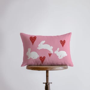 May include: A pink rectangular pillow featuring white rabbit silhouettes and red hearts with white polka dots. The pillow is resting on a wooden stool. The design is likely for Easter or a spring theme.