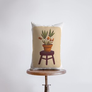 May include: A throw pillow with a beige background featuring a hand-painted illustration of a potted plant with orange flowers on a small wooden stool.