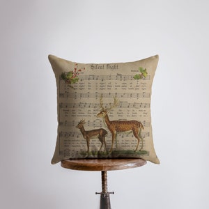 Puede incluir: Una almohada beige con un diseño de partitura de música vintage que presenta la letra del villancico "Noche de Paz". Dos ciervos están representados en la partitura, uno de pie y otro acostado. La almohada está sentada en un taburete de madera.