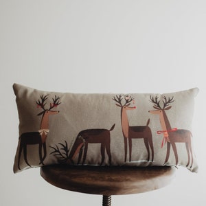 Puede incluir: Una almohada lumbar beige con un diseño festivo que presenta cuatro renos estilizados con acentos rojos. Los renos se representan en un estilo juguetón y caprichoso.