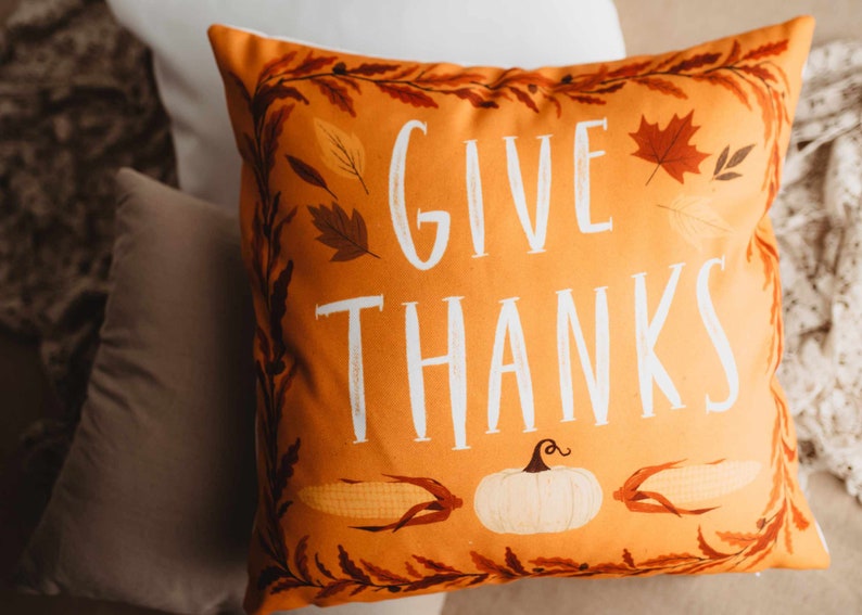 Pu&ograve; includere: Cuscino arancione con il testo "Give Thanks" in lettere bianche. Il cuscino ha un tema autunnale con illustrazioni di foglie, mais e una zucca.