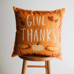 Pu&ograve; includere: Cuscino da lancio arancione con il testo "GIVE THANKS" in lettere bianche. Il cuscino presenta una zucca, mais e foglie autunnali.