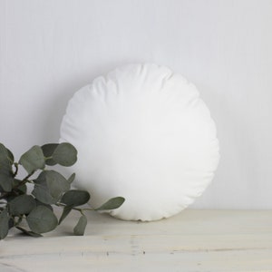 May include: A circular white cushion with a soft, textured finish. It rests on a white wooden surface, with a sprig of green eucalyptus leaves in the foreground. The cushion is a neutral colour and could be used as a decorative item.
