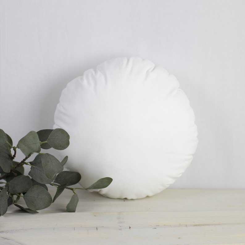 May include: A circular white cushion with a soft, textured finish. It rests on a white wooden surface, with a sprig of green eucalyptus leaves in the foreground. The cushion is a neutral colour and could be used as a decorative item.