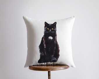 May include: A white pillow with a black cat illustration. The cat is sitting with its front paws tucked in and looking directly at the viewer. The cat has a white patch on its chest.