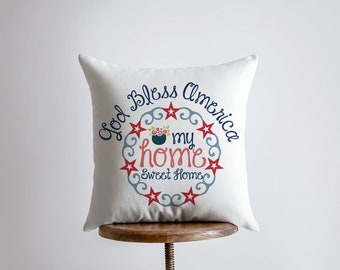 Op de afbeelding: Wit kussen met een rood, wit en blauw ontwerp met de tekst "God Bless America my home Sweet Home".