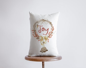 May include: White throw pillow with a brown and red wreath design and the word "Joy" in red script. The wreath is decorated with small bells and a yellow bow.