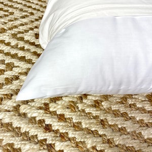 May include: A white pillow on a woven rug with a diamond pattern. The rug is made of natural fibers and has a light brown and cream color.