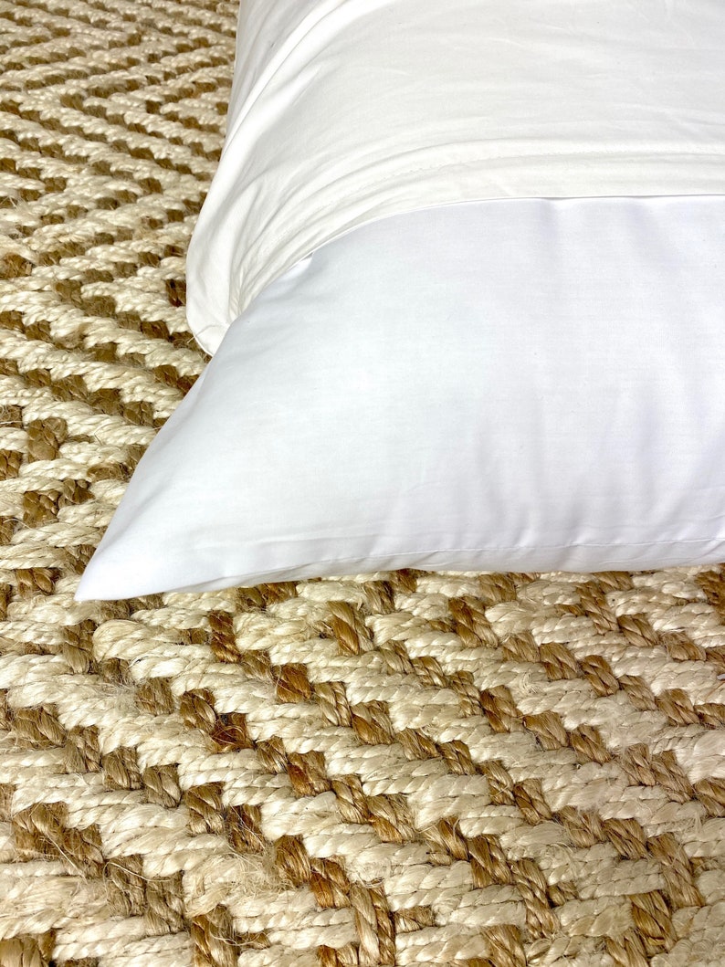May include: A white pillow on a woven rug with a diamond pattern. The rug is made of natural fibers and has a light brown and cream color.