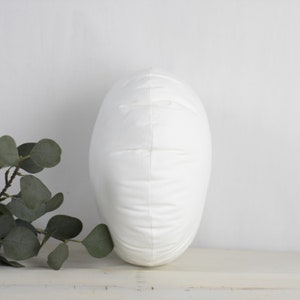 May include: A white, oval cushion with a soft, textured surface. The cushion is upright, displaying its shape and design. The background is a neutral white, emphasising the cushion's clean aesthetic.