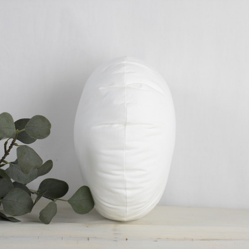 May include: A white, oval cushion with a soft, textured surface. The cushion is upright, displaying its shape and design. The background is a neutral white, emphasising the cushion's clean aesthetic.