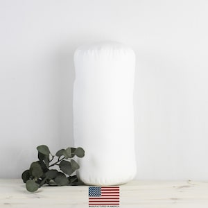 Puede incluir: Almohada cilíndrica blanca con una funda de tela lisa. La almohada está sobre una superficie de madera clara con una rama de hojas verdes delante. El texto "MANUFACTURED IN AMERICA" está impreso debajo de una pequeña bandera estadounidense.
