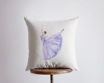 May include: A square, off-white throw pillow featuring a watercolor illustration of a ballerina in a flowing lavender dress. The dancer is poised mid-leap, with one leg extended and arms gracefully raised. The pillow is displayed on a wooden stool.