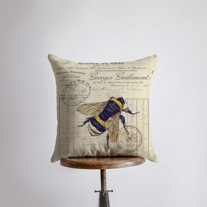 May include: A beige pillow with a vintage French design featuring a bee and floral accents. The pillow has a text overlay that reads "Maison de Gros" and "Georges Guillemont".