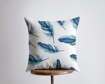 Puede incluir: Una almohada blanca con un patrón de plumas azules en acuarela. La almohada está sentada en un taburete de madera.