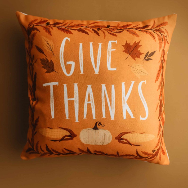 Pu&ograve; includere: Cuscino arancione con bordo bianco e il testo "GIVE THANKS" in lettere bianche. Il cuscino presenta un design autunnale con foglie, mais e una zucca.