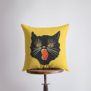 Pode incluir: Uma almofada quadrada amarela com um desenho de rosto de gato preto. O gato tem olhos amarelos, bigodes pretos e uma língua vermelha. A almofada está colocada sobre um banco de madeira. O design tem uma estética vintage.