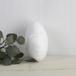 May include: A white, oval cushion with a smooth, slightly textured surface. The cushion is standing upright, displaying its shape. A sprig of green eucalyptus leaves is visible on the left, adding a natural element to the composition.