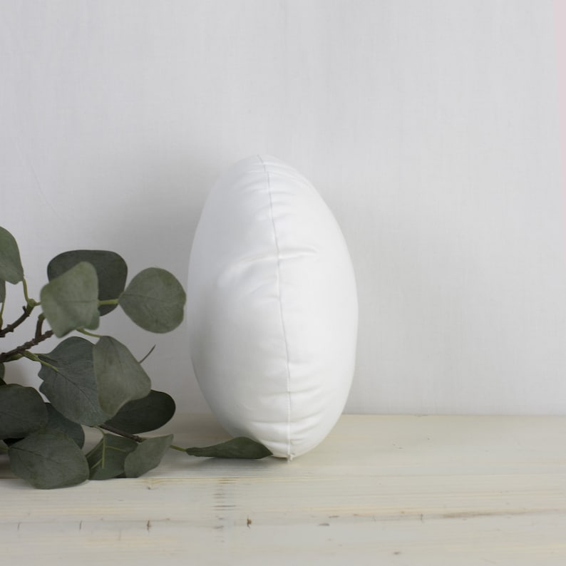 May include: A white, oval cushion with a smooth, slightly textured surface. The cushion is standing upright, displaying its shape. A sprig of green eucalyptus leaves is visible on the left, adding a natural element to the composition.