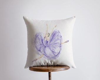 May include: A square, off-white throw pillow featuring a watercolor illustration of a ballerina in a dynamic pose. The ballerina is depicted in shades of purple, with a flowing tutu and arms outstretched. The pillow is resting on a wooden stool.