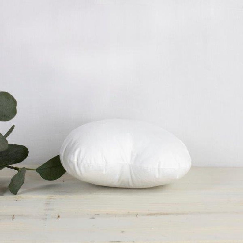 May include: A white round pillow with a smooth surface. The pillow is sitting on a light wood surface.