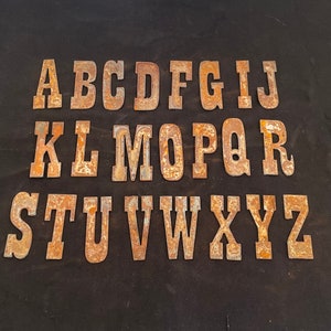 May include: A set of rusty metal alphabet letters, in uppercase, arranged in alphabetical order. The letters are painted a light brown color and have a worn, vintage look.