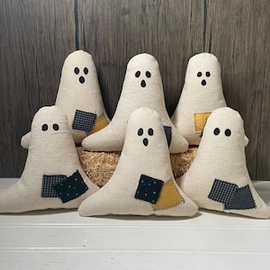 May include: Six handmade ghost decorations. Each ghost is made of off-white fabric with black button eyes and a stitched mouth. Patches of blue, black, and yellow fabric are sewn onto the bottom. The ghosts are arranged on a bed of straw.