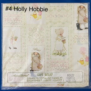 May include: A sheet of gift wrap featuring a Holly Hobbie design. The wrap is white with a green and pink floral pattern. The design includes a girl with a watering can, a cat, and the text "Holly Hobbie".