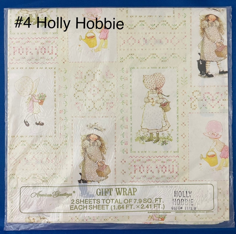 May include: A sheet of gift wrap featuring a Holly Hobbie design. The wrap is white with a green and pink floral pattern. The design includes a girl with a watering can, a cat, and the text "Holly Hobbie".