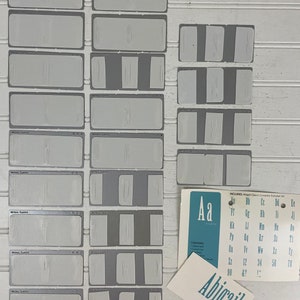 May include: A set of white plastic stencils for lettering, featuring the complete alphabet in uppercase and lowercase letters, numbers, and punctuation marks. The stencils are arranged in rows on a white surface. The word "Abigail" is written in blue letters on a white card.