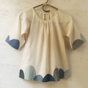 Puede incluir: Un vestido color crema de manga corta con cuello redondo y dobladillo festoneado. El dobladillo y los puños de las mangas presentan un patrón de ondas azul, gris y azul oscuro. El vestido está colgado de una percha de madera sobre un fondo neutro.
