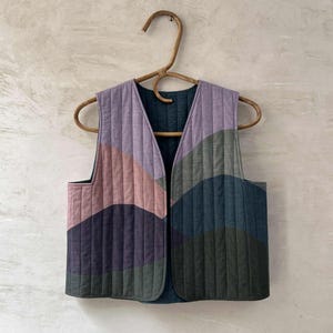 May include: A sleeveless quilted vest featuring a vertical stitched pattern and a color-blocked design in shades of lavender, dusty pink, dark purple, olive green, and teal blue, hanging on a curved wooden hanger with an open front design.