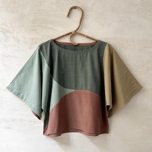 May include: A short-sleeved top featuring a colour-blocked design in shades of sage green, dark forest green, terracotta, and mustard yellow. The garment has a wide, boxy cut with a round neckline and is displayed on a curved wooden hanger. Text reads FOREST / RED EARTH.