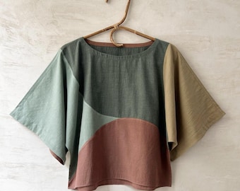 May include: A short-sleeved top featuring a colour-blocked design in shades of sage green, dark forest green, terracotta, and mustard yellow. The garment has a wide, boxy cut with a round neckline and is displayed on a curved wooden hanger. Text reads FOREST / RED EARTH.