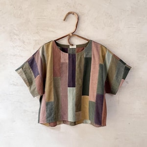 Może przedstawiać: Krótka bluzka z patchworkowym wzorem. Bluzka posiada pionowe prostokątne panele w odcieniach fioletu, różu, zieleni i beżu. Bluzka wisi na drewnianym wieszaku na neutralnym tle.
