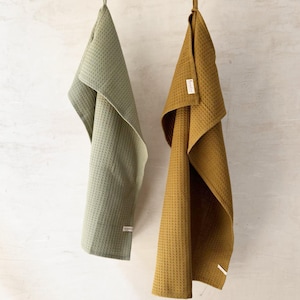 May include: Two kitchen towels hanging on a clothesline. The towel on the left is sage green and the towel on the right is brass yellow.