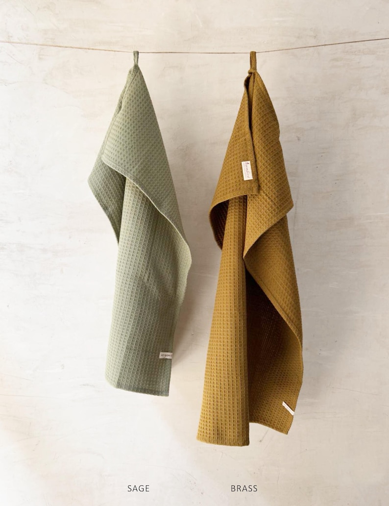 May include: Two kitchen towels hanging on a clothesline. The towel on the left is sage green and the towel on the right is brass yellow.