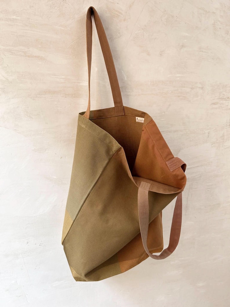 K&ouml;nnte beinhalten: Eine Tragetasche aus Segeltuch in Olivgr&uuml;n und Braun mit langen, braunen Tr&auml;gern. Die Tasche ist offen und zeigt das Innere. Die Tasche hat ein schlichtes, minimalistisches Design, das sich zum Tragen von Lebensmitteln oder Alltagsgegenst&auml;nden eignet.