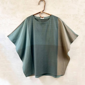 May include: A teal, boxy top with a round neckline and short sleeves, hanging on a wooden hanger. The top features a colour block design with vertical panels of teal, urban blue, and camel. The text "PETROL / URBAN BLUE / CAMEL" is at the bottom.