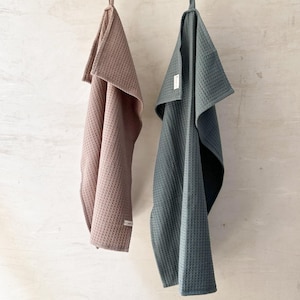May include: Two waffle weave kitchen towels hanging on a string. The towel on the left is a light brown color and says "GREY TAUPE". The towel on the right is a dark green color and says "STORM".