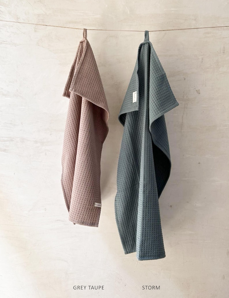 May include: Two waffle weave kitchen towels hanging on a string. The towel on the left is a light brown color and says "GREY TAUPE". The towel on the right is a dark green color and says "STORM".