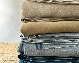 May include: A stack of folded textiles in various neutral colors. The stack includes tan, beige, and blue fabrics. The fabrics appear to be linen or a similar natural material, with visible textures and folds. The arrangement suggests bedding or home decor items.