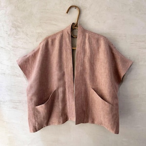 May include: A dusty rose linen jacket with short sleeves and two front pockets. The open-front design is displayed on a wooden hanger against a neutral wall. The jacket's texture is visible, suggesting a breathable fabric, ideal for warmer weather.