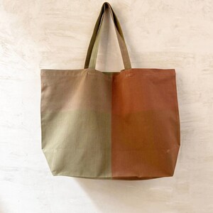 K&ouml;nnte beinhalten: Eine gro&szlig;e Tragetasche mit zweifarbigem Design. Eine Seite ist salbeigr&uuml;n, die andere erdfarbenes Rosa. Die Tasche hat passende salbeigr&uuml;ne Griffe und den Text "SAGE / EARTH PINK" unten.