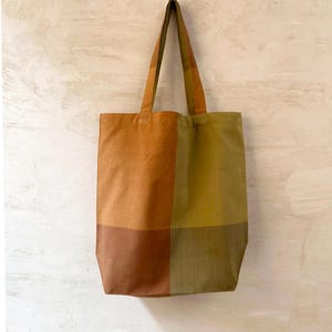 K&ouml;nnte beinhalten: Eine rechteckige Tragetasche mit einem Patchwork-Design in Rost-, Oliven- und Braunt&ouml;nen. Die Tasche hat lange Henkel und der Text "RUST / OLIVE" ist unten aufgedruckt.