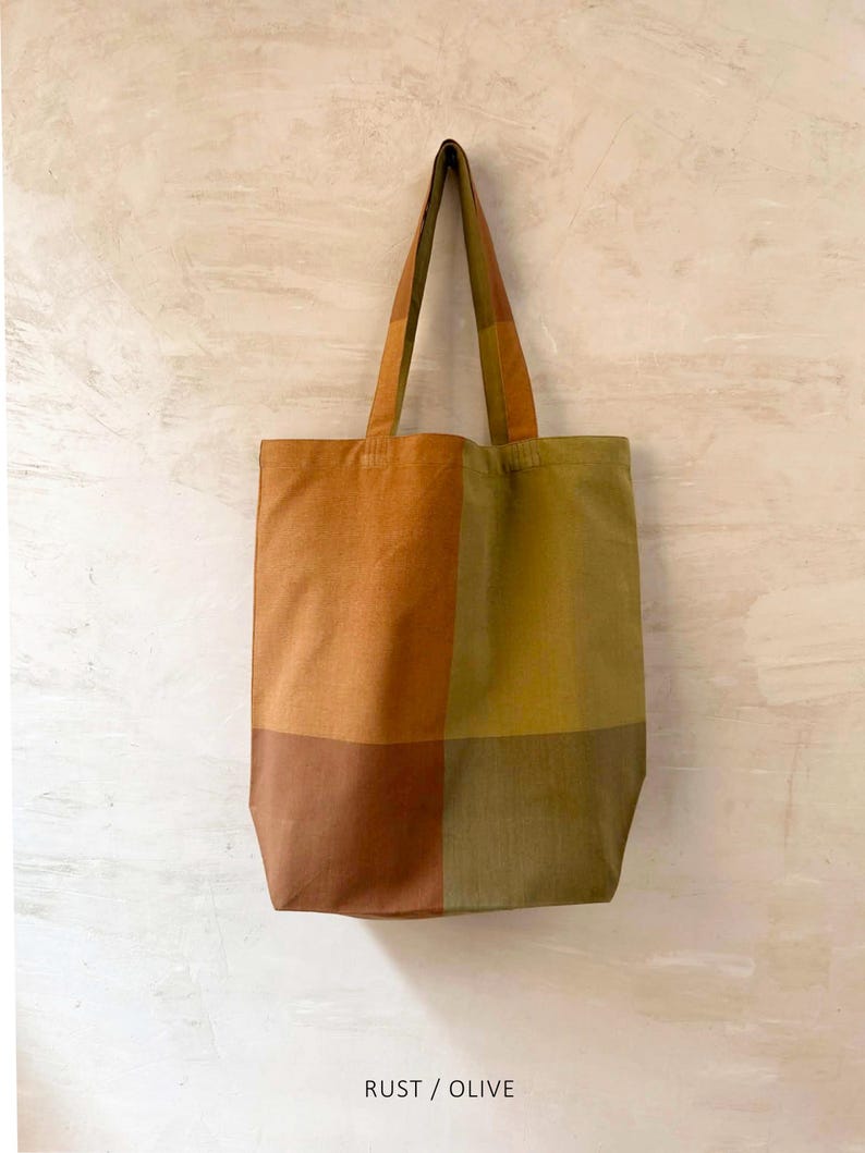 K&ouml;nnte beinhalten: Eine rechteckige Tragetasche mit einem Patchwork-Design in Rost-, Oliven- und Braunt&ouml;nen. Die Tasche hat lange Henkel und der Text "RUST / OLIVE" ist unten aufgedruckt.