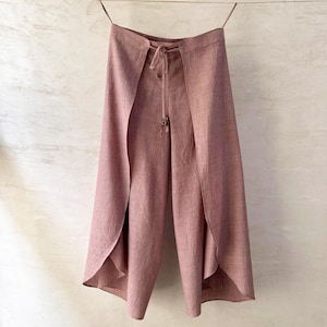 May include: Dusty rose linen trousers with a distinctive wrap-around design. The trousers have a drawstring waist and a flowing, wide-leg silhouette. The fabric looks lightweight. The trousers are hanging from a thin string. The measurements are in centimeters.