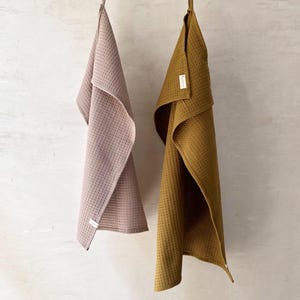 May include: Two waffle weave kitchen towels hanging on a clothesline. The towel on the left is grey taupe and the towel on the right is brass.