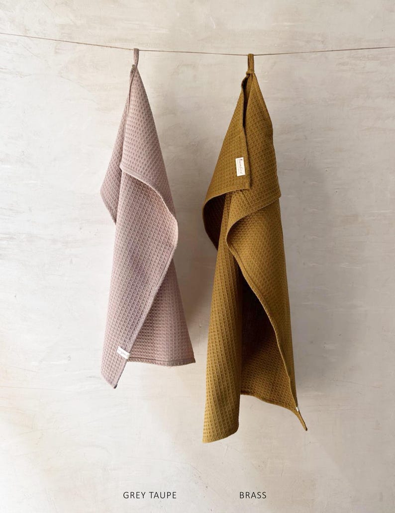 May include: Two waffle weave kitchen towels hanging on a clothesline. The towel on the left is grey taupe and the towel on the right is brass.